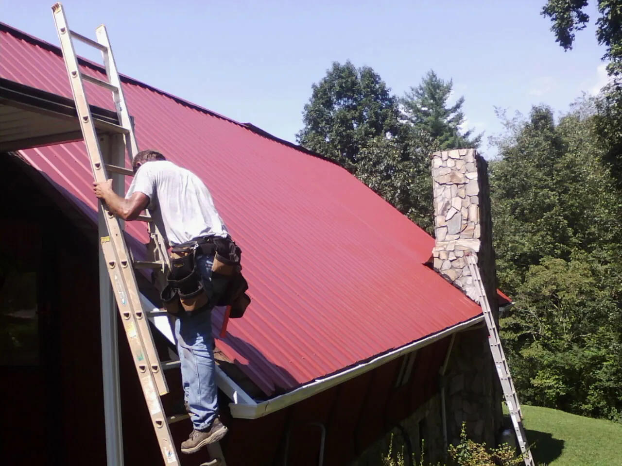 Roofer on a ladder beside a red metal roof and stone chimney — Soffit & Fascia Repair by Roofing Brothers in Santa Rosa Beach
